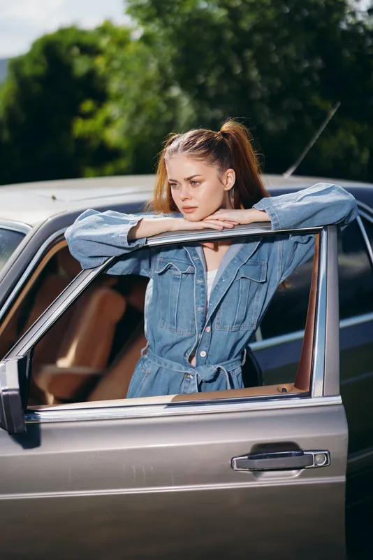 Stylish young woman in denim jumpsuit posing by a vintage car, exuding confidence and casual elegance in a beautiful outdoor setting.