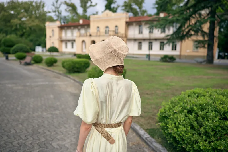 Vintage dress and bonnet woman seen from behind walking along a garden path toward a historical manor, creating a timeless fashion scene in an outdoor setting, tranquil