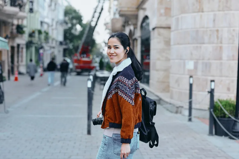 Woman with backpack and camera on a city street, wearing a patterned brown jacket, denim jeans, and a white scarf, smiling as she walks past historic buildings and modern storefronts frame the scene.