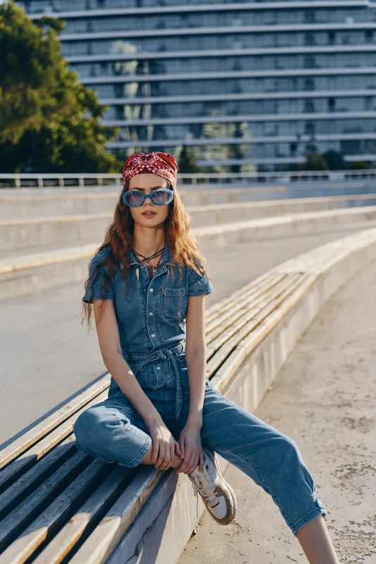 Young woman wearing denim outfit and red headscarf sitting outdoors on wooden bench with sunglasses under bright sunlight in urban environment.