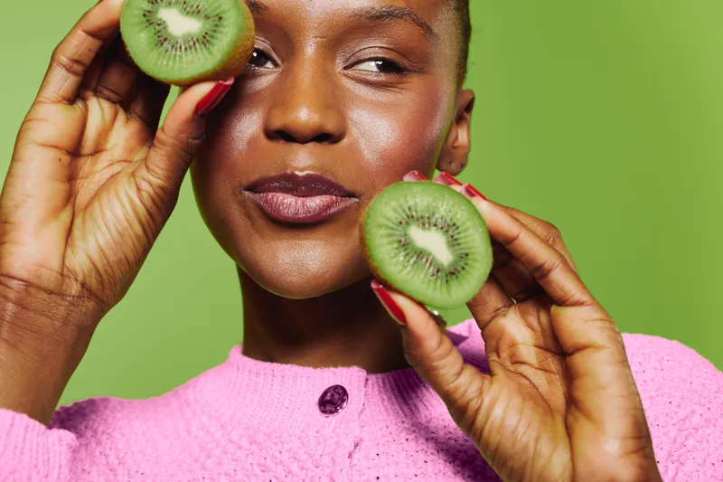Close-up of a smiling dark skinned African woman holding two halves of a kiwi fruit near her face. Healthy lifestyle, fresh fruit, and natural beauty on green background concept.