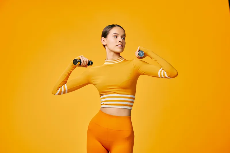 Woman fitness, sport: Fit young woman exercising with handheld dumbbells in a bright studio, wearing matching orange activewear, confident posture and strength training lifestyle visual.