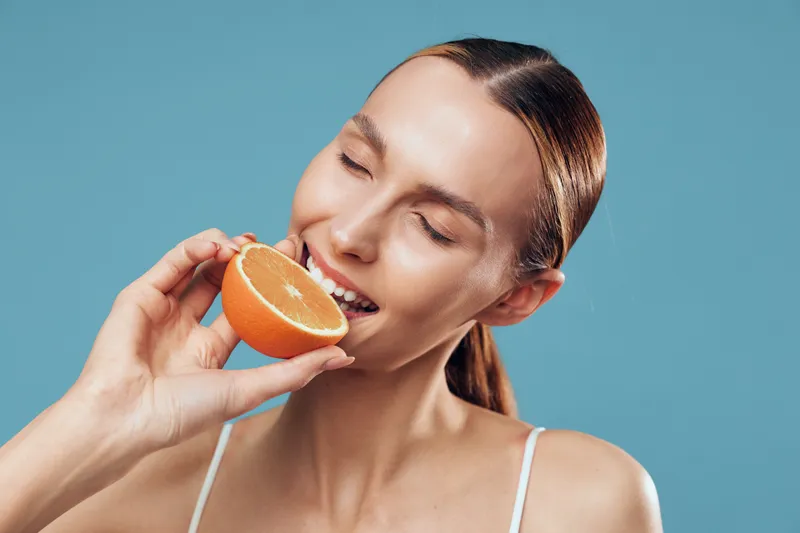 Young woman with closed eyes enjoying fresh orange slice, natural skincare, healthy lifestyle, bright blue background, soft lighting, closeup portrait, casual white top