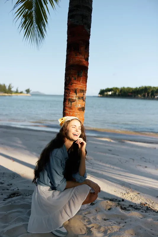 Young woman sits beside a palm tree on a sandy beach, smiling softly as warm sun lights her features. Calm water and blue sky create a peaceful, sunlit moment.