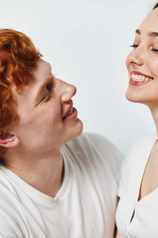 Couple smiling and looking at each other with joy and affection on white background. Young adults in casual clothing sharing moment of happiness and connection.