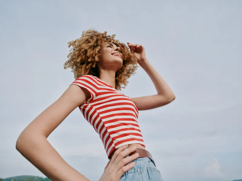 Joyful young woman with curly hair stands outdoors in a red and white striped tee and denim shorts, smiling confidently as she looks toward the bright sky.