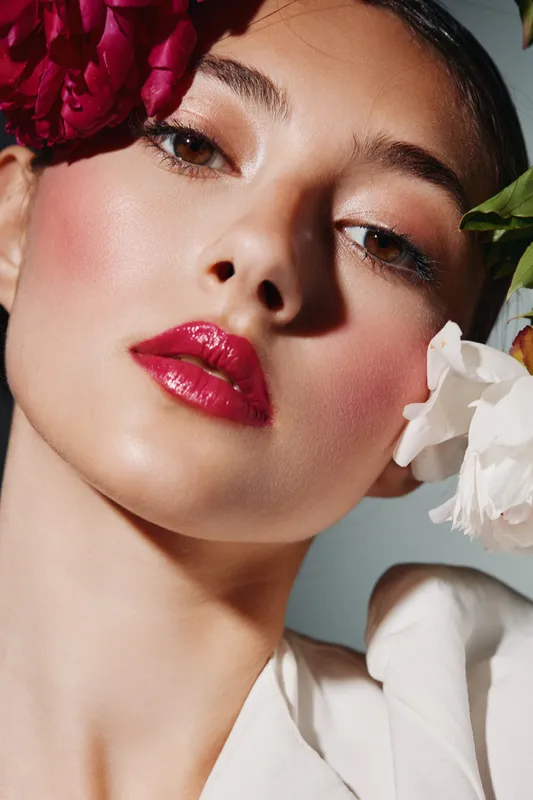 Close-up of a young woman with glossy red lips and dewy skin wearing vibrant makeup surrounded by fresh flowers. Concept of beauty, fashion, and creative portrait photography.