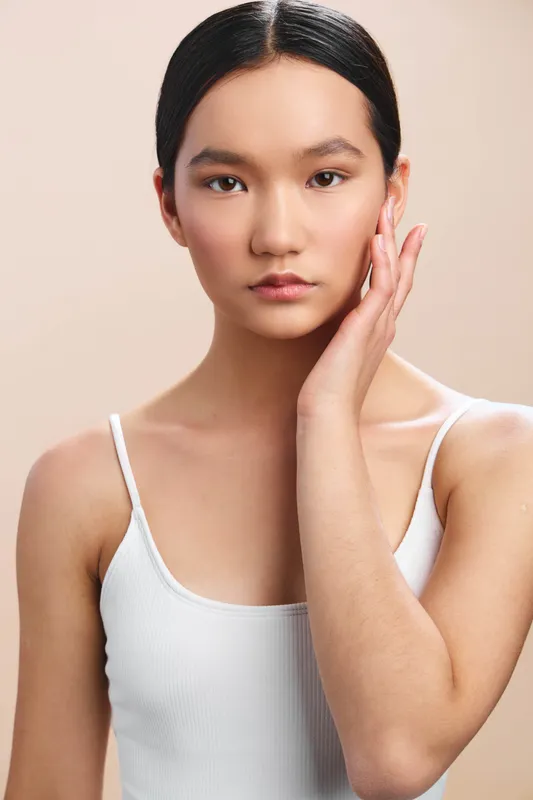Asian woman with dark hair styled sleekly, wearing a white sleeveless top, softly touching her face, looking at the camera with a calm, contemplative expression suitable for beauty and lifestyle