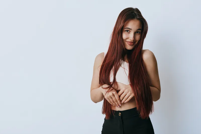 Young woman with long red hair posing and smiling against light background. Casual outfit with black pants and white top, natural beauty and confident expression in studio.