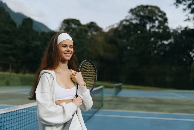 Smiling young woman in sporty attire holding a tennis racket on a sunny day at a tennis court, evoking feelings of joy, health, and active lifestyle