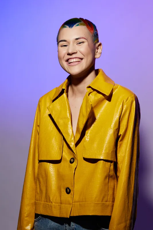 young woman with colorful short hair smiling joyfully in a bright yellow jacket against a purple background in a modern studio setting