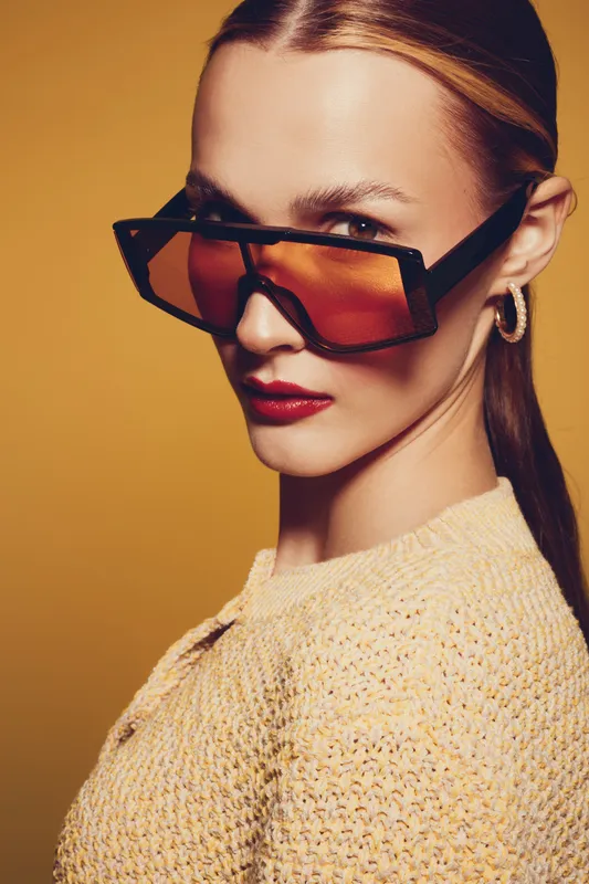 Stylish woman with slicked hair wears oversized orange sunglasses and textured yellow sweater. Close-up portrait on warm yellow background. Concept of fashion, confidence, and modern style.