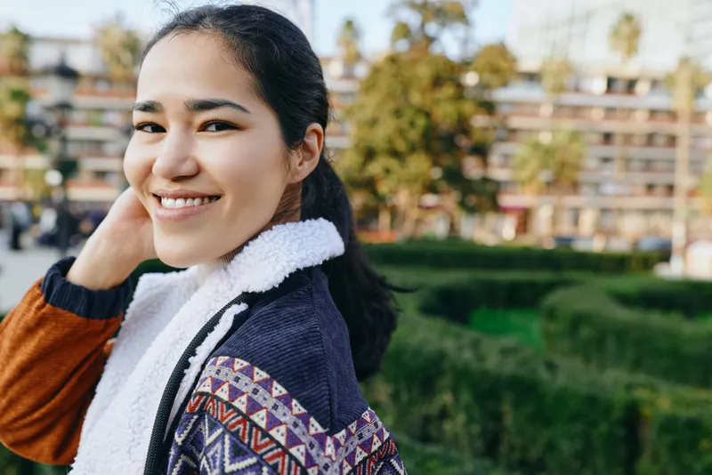 Woman with a warm smile outdoors, wearing a patterned sweater and scarf, in a sunny park setting with greenery and buildings in the background
