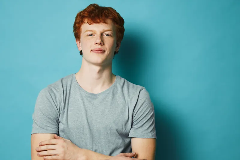 Young man with red hair and freckles wearing gray t shirt standing with arms crossed against blue background. Casual confident expression with simple studio portrait lighting.