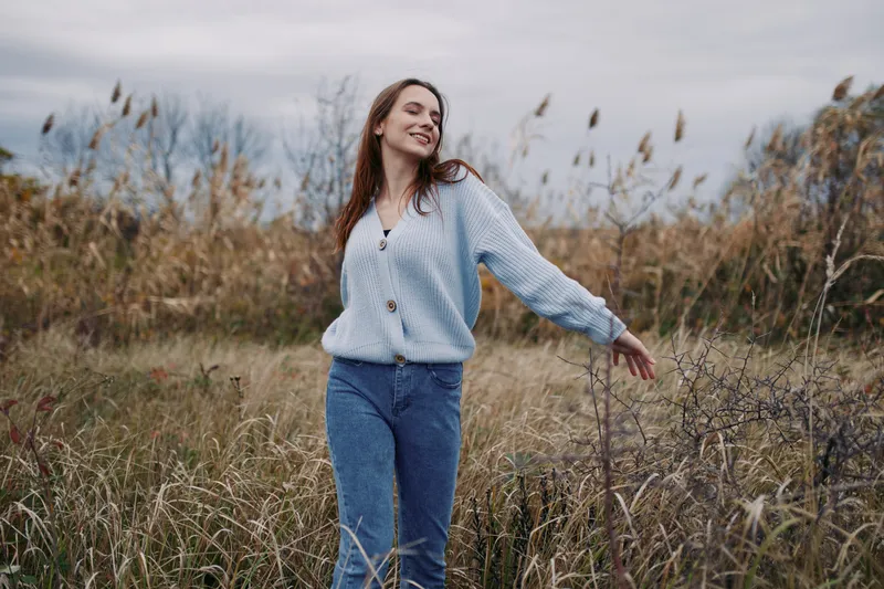 A candid portrait of a young woman in a breezy field, smiling softly as she moves freely, showcasing natural posture and genuine emotion under muted skies.