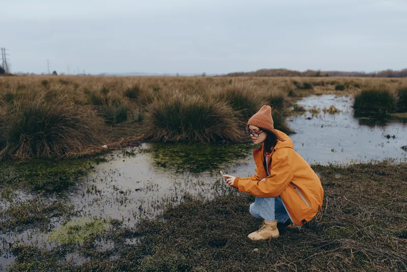 Woman lifestyle outdoors, kneeling by wetland water, wearing knitted hat and jacket, tall grass in autumn scenery, peaceful nature walk, soft overcast light