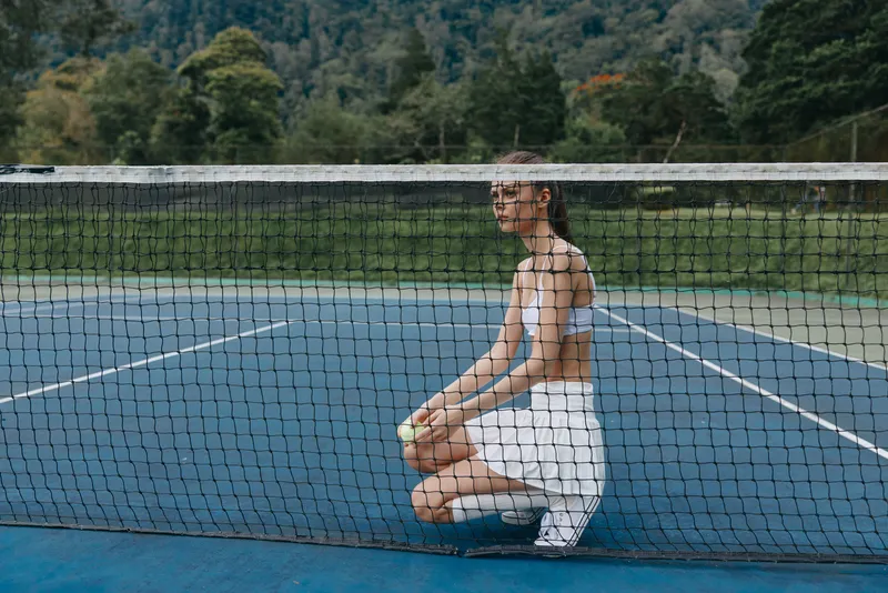 Young female tennis player crouching on court, showcasing athletic wear and determination amidst a lush green background