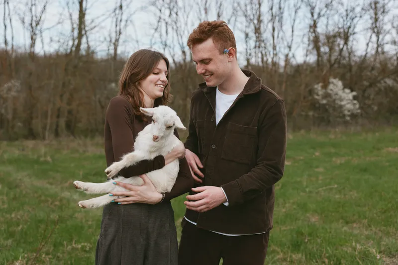Joyful couple outdoors with a fluffy goat kid in a grassy field, warm light, candid lifestyle moment, affection and play, natural setting conveying rural charm, gentle bonding between people and