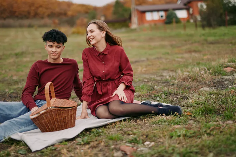 A candid outdoor moment captures two people sharing a simple picnic on a grassy field, genuine smiles, natural light, relaxed posture, and an authentic, wholesome vibe.