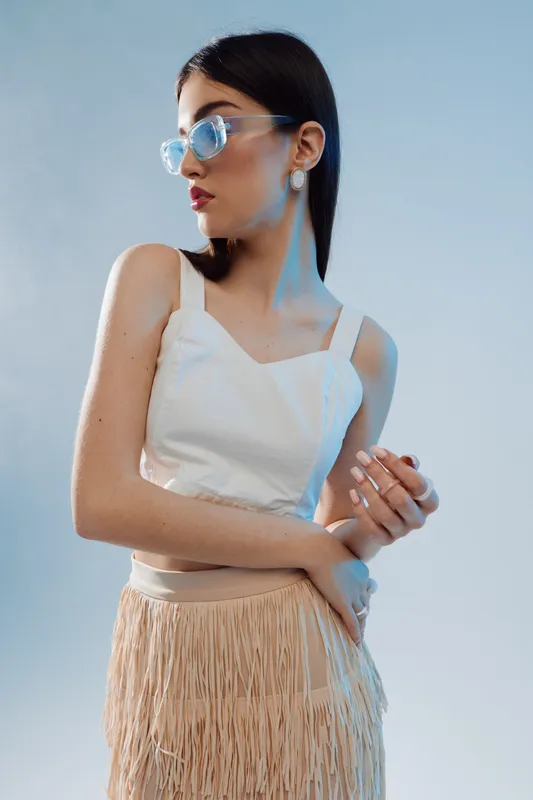 Young woman in white top and beige fringed skirt poses wearing transparent sunglasses. Studio shot with soft lighting on a neutral background. Concept of woman beauty fashion style.