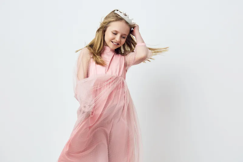 Cheerful young girl in a pink dress, playing with her hair, showcasing happiness and innocence against a soft white background