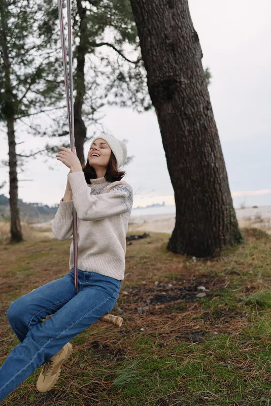 Girl on a swing in an outdoor nature setting with trees and autumn ground, smiling and relaxed in a sweater and beanie, capturing a carefree joyful moment in a park