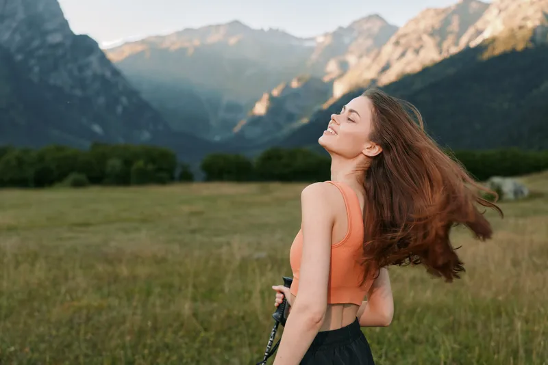 A cheerful woman stands in a wide open field with mountains in the distance, wearing an orange top. Her eyes are closed, head tilted up, and hair flowing as she smiles.