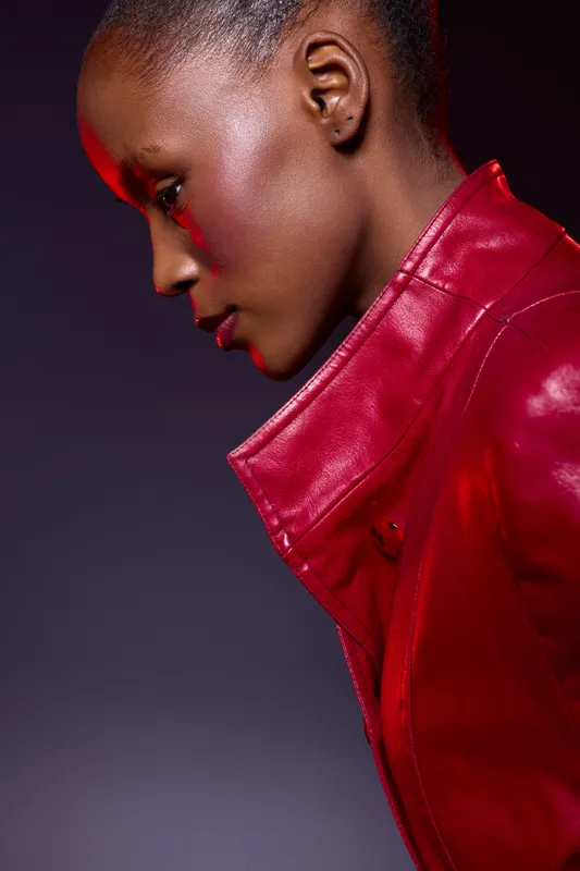 Profile of a young black woman wearing a red leather jacket. She has dark skin and short hair, posing against a dark purple background. Concept of fashion, style, and confidence.