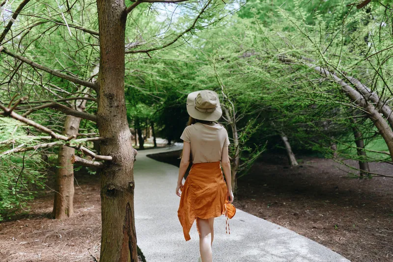 Woman walking on forest path wearing a sun hat and orange skirt, back view on a gravel trail surrounded by trees and green foliage for outdoor nature stroll.