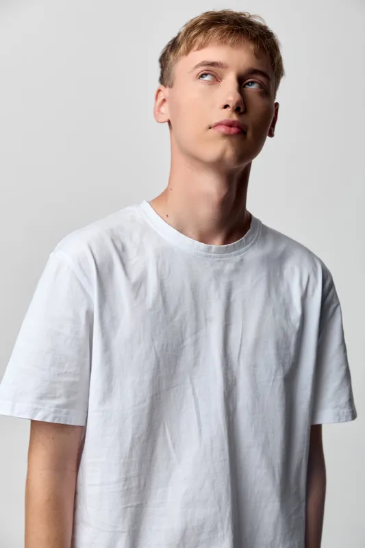 Youth, male in white t shirt looking upward against a plain studio backdrop, thoughtful expression and minimal fashion mood for lifestyle and advertising use