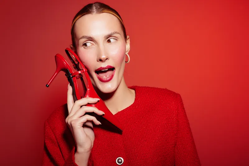 Young woman in red sweater holds red high heel shoe near face, looking sideways with playful expression. Bright red background creates bold and vibrant fashion concept.