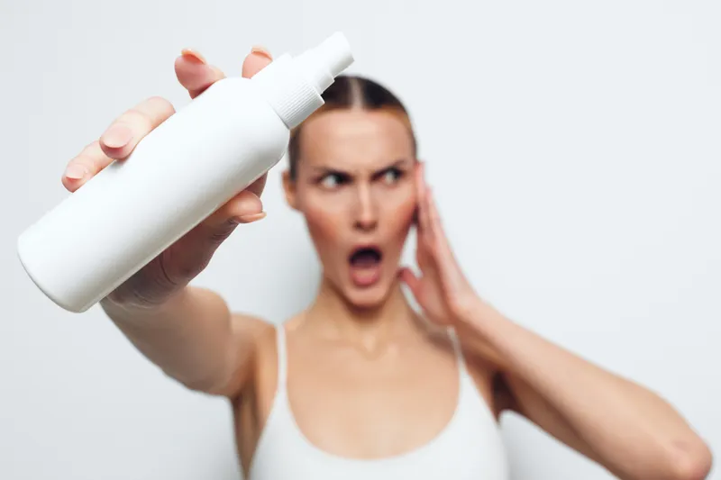 A surprised woman in a white tank top holds a white spray bottle close to the camera with one hand while touching her face with the other. Concept of skincare and product reaction.