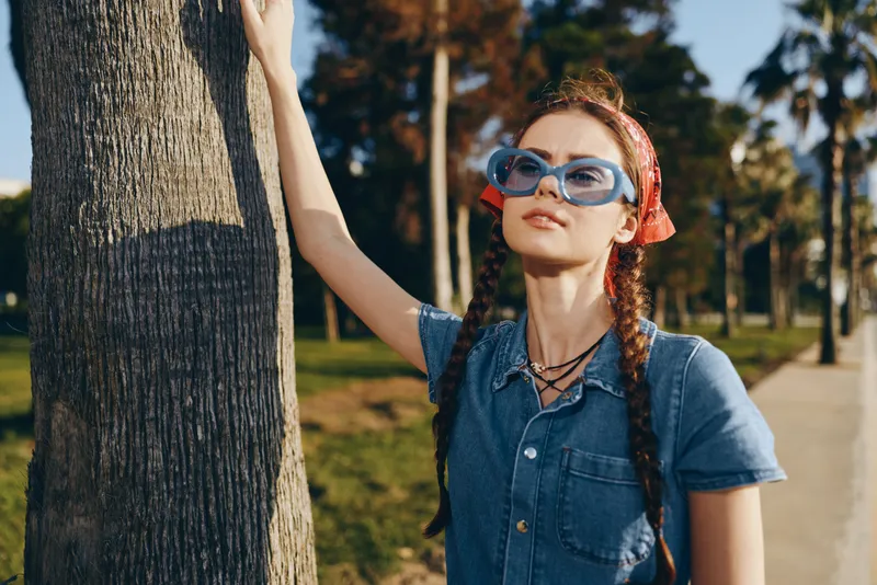 Lifestyle woman in modern boho-western denim street style poses outdoors wearing blue sunglasses and a red headscarf with film color warm tones.