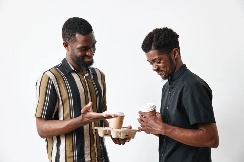 Two African American men with dark skin holding coffee cups and smiling, casual clothing, friendly interaction, isolated on white background.