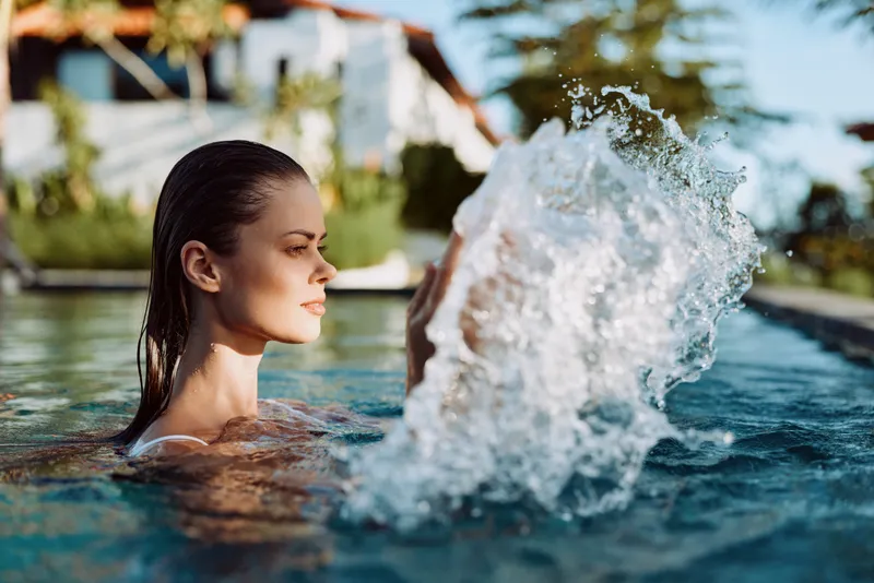 Cheerful woman in a pool, water splash captured mid motion, sunny outdoor scene, relaxed posture, warm breeze, natural light, vivid blue water, lush background, lifestyle moment