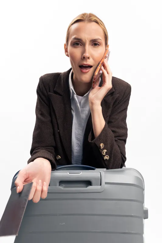 Stressed young woman in brown blazer holding suitcase talks on phone with worried expression. Concept of travel problems, delay, and communication issues during trip preparation.