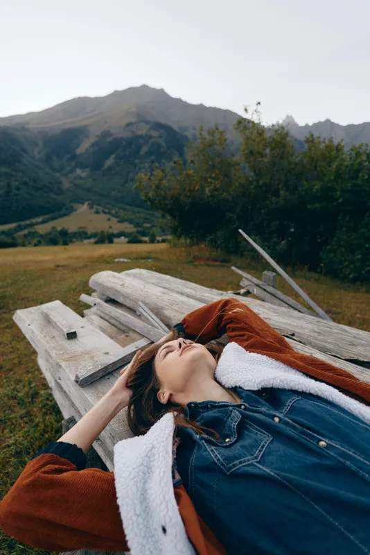 Woman lying on a wooden picnic table in mountain nature outdoors, relaxation and landscape scene with meadow and trees, portrait of calm travel and peaceful escape feeling.