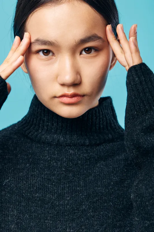 Young woman with clear skin and dark hair wearing black sweater touching face with fingers, looking serious on blue background. Closeup portrait of thoughtful female with natural expression.