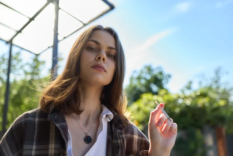 A casual outdoor portrait of a confident woman smoking a cigarette in daylight, wearing a plaid shirt, in a sunny garden. A lifestyle moment captured with simplicity and ease