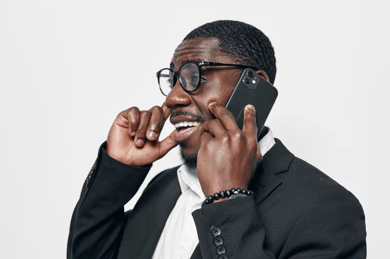 Black man with dark skin and glasses talking on smartphone and smiling in formal suit and white shirt. Business communication, technology and positive expression in office or studio.