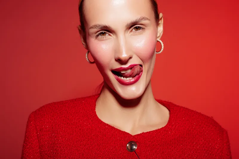 Young woman in red sweater poses with tongue out on red background. Close-up portrait with hoop earrings and playful expression. Concept of fun, fashion, and bold style.