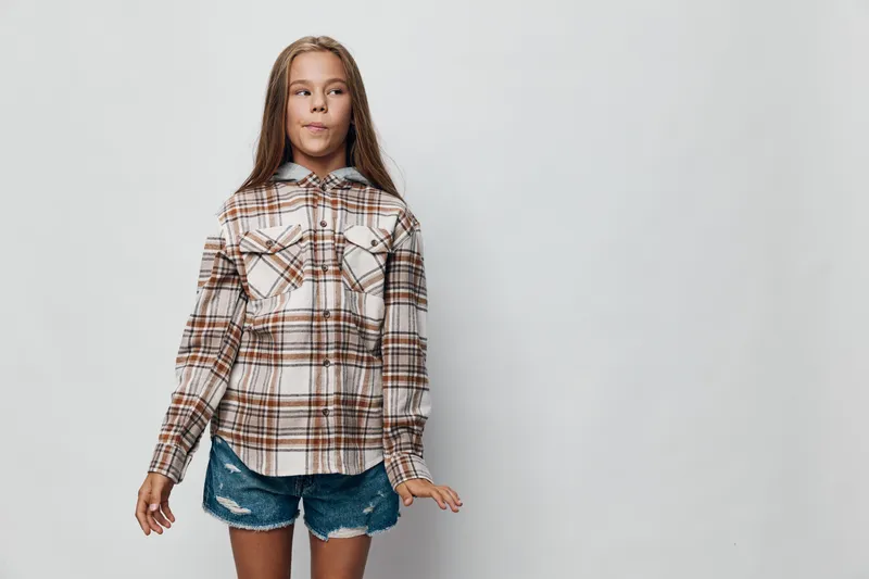 Girl standing wearing plaid shirt and denim shorts with long hair looking sideways on light background. Casual style, childhood, fashion and youth with confident pose.