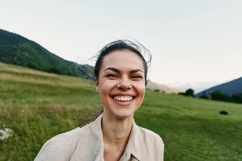 A young woman with brunette hair smiles broadly in an open countryside field, capturing a carefree moment in natural light, casual attire, and a warm afternoon mood for lifestyle and wellness