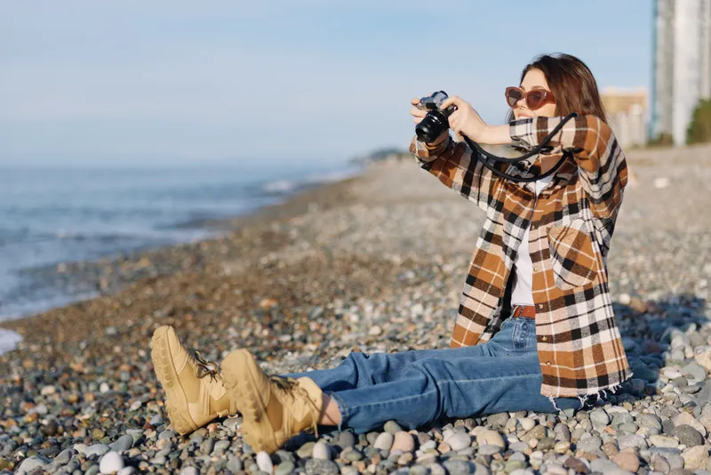 Beach photography by a photographer on a pebbled coast, outdoors lifestyle with a woman in a plaid jacket, casually posing and capturing the sea with a handheld camera amid bright daylight.