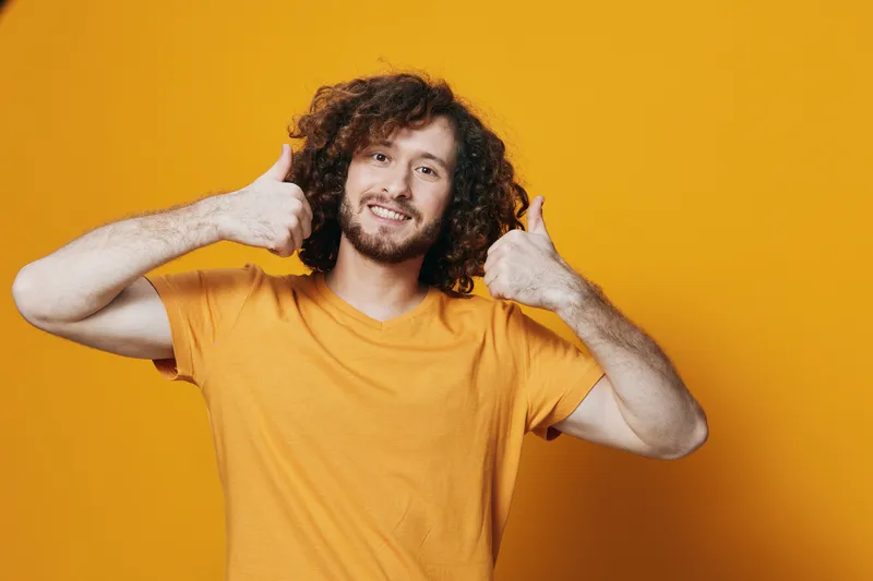 Cheerful young man with curly hair in a bright orange t shirt, giving thumbs up in front of a vibrant yellow background, embodying positivity and joy