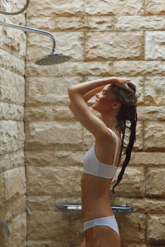 young woman showering outdoors under rainfall, wearing white underwear, wet hair, stone wall background, natural light, serene mood