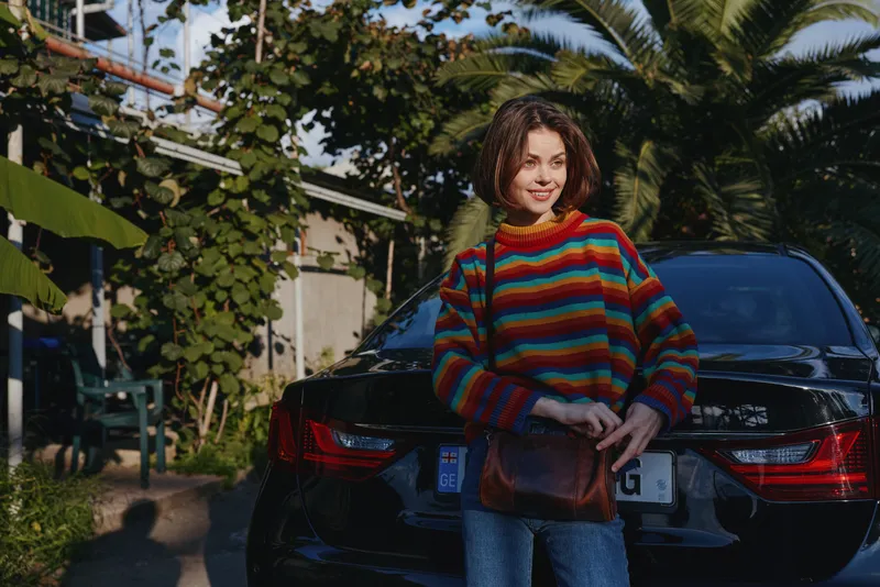 Young woman in colorful sweater standing by black car with leather bag, palm trees and sunlight in background. Outdoor portrait, casual fashion, travel and city lifestyle.