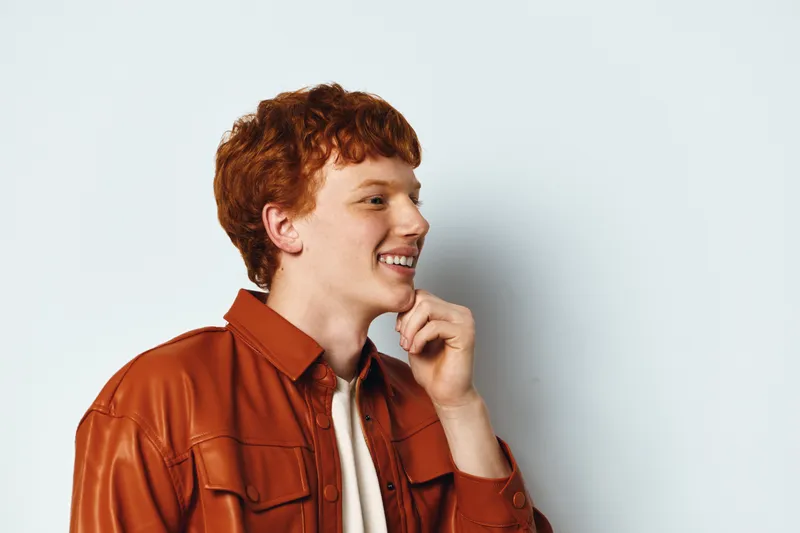 Young man with red hair smiling in orange jacket against white background. Casual style, side profile, friendly expression and thoughtful pose with hand near chin.
