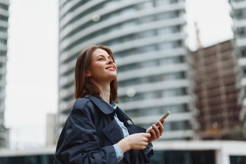 A smiling young woman in a navy jacket gazes upward while holding a smartphone in a modern city setting, conveying confidence, curiosity, and urban exploration.