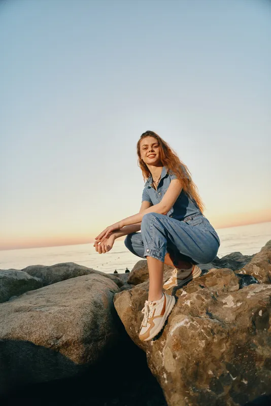 Lifestyle woman in modern boho-western denim street style sitting on rocks near ocean during golden hour with relaxed smile and warm film color tones.
